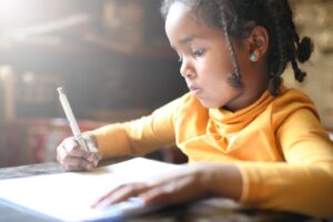 A young girl in a yellow shirt focuses intently on writing with a pencil in a notebook. She has braided hair and the setting is warmly lit.